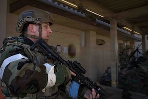 An Airman with a camouflage green and black protective coat holds a black rifle aiming down while looking forward. He wears a tan helmet that exposes his serous but calm expression on his face.