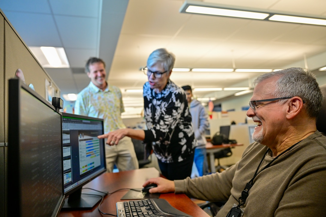 A man sits at a computer and a woman stands next to him, pointing at the screen