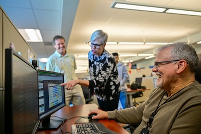 A man sits at a computer and a woman stands next to him, pointing at the screen