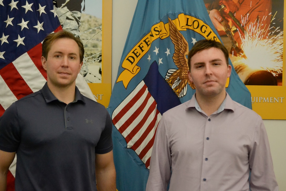 Two men stand in front of the American and Defense Logistics Agency flags