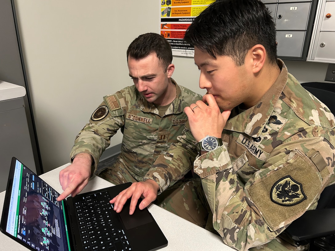 Two men in military uniforms sit in front of a laptop