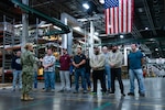 A sailor in uniform stands in front of a group of civilians in a warehouse