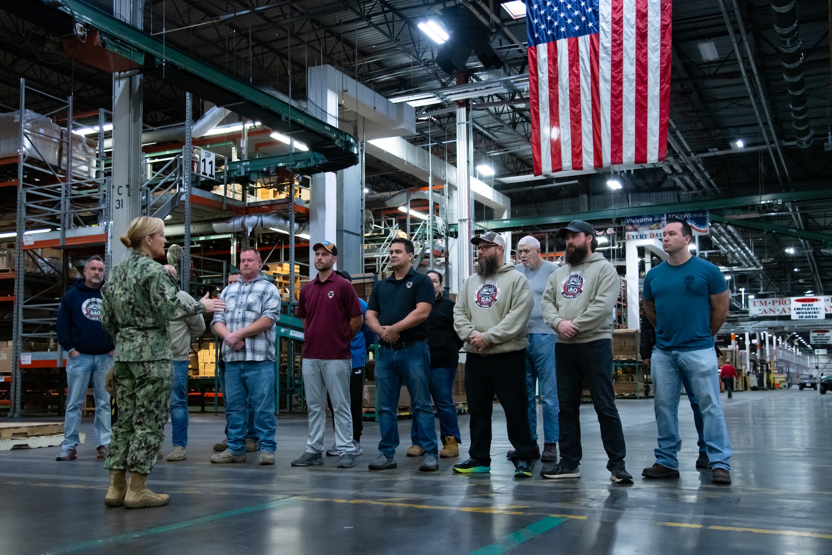 A sailor in uniform stands in front of a group of civilians in a warehouse