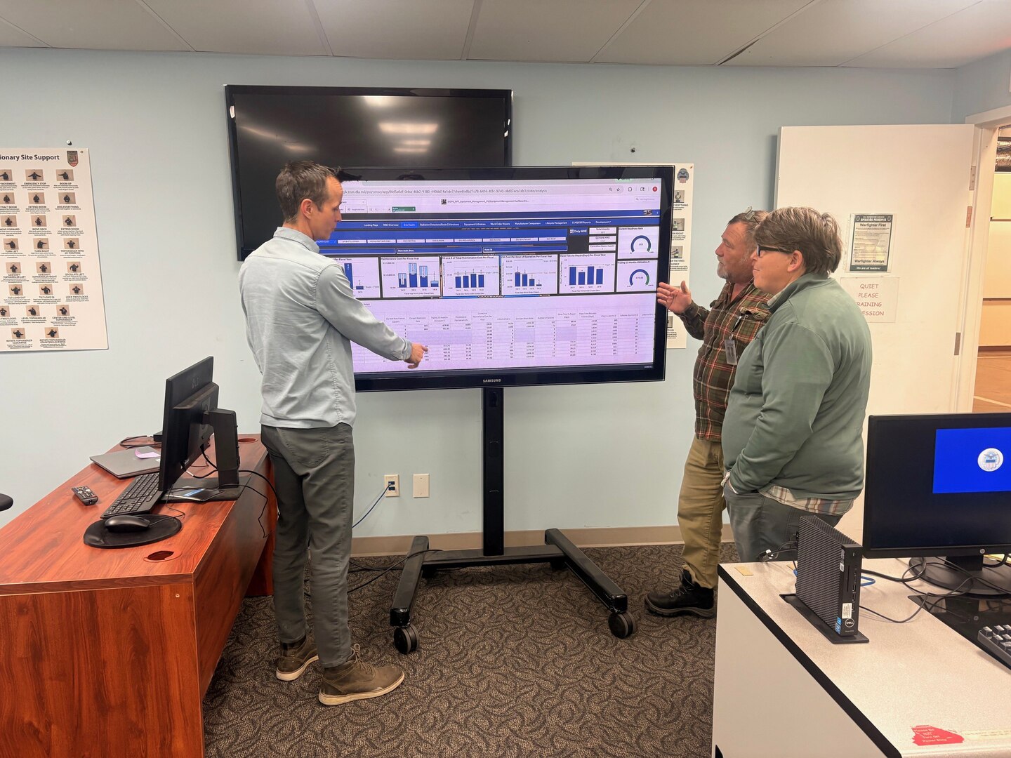 Three men stand around a monitor displaying a dashboard