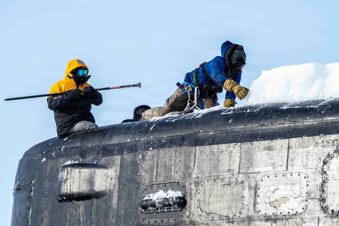 Two sailors, both in winter gear and one with a tool, remove ice from the sail of a submarine against a blue sky.