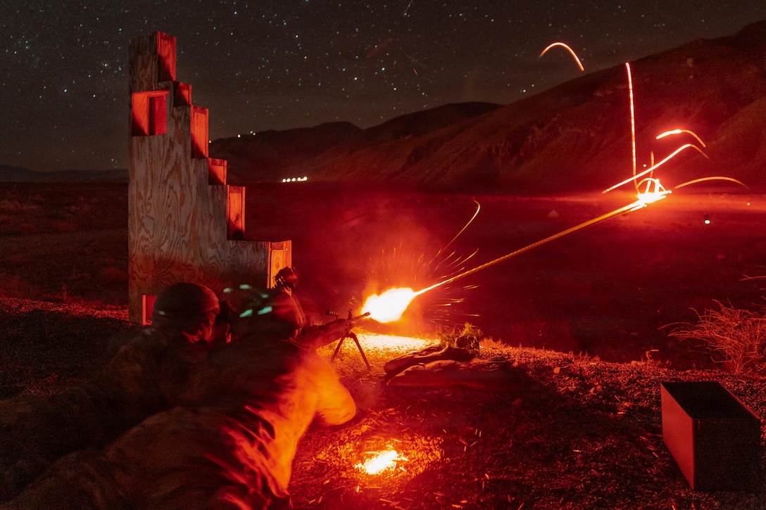 Two soldiers lying on the ground fire a weapon, creating red laser streaks across a field and mountain under a starry night sky.