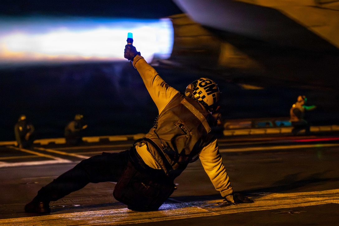 A sailor raises a baton while leaning on the ground as an aircraft launches in the dark aboard a ship, creating a bluish, orange blast.