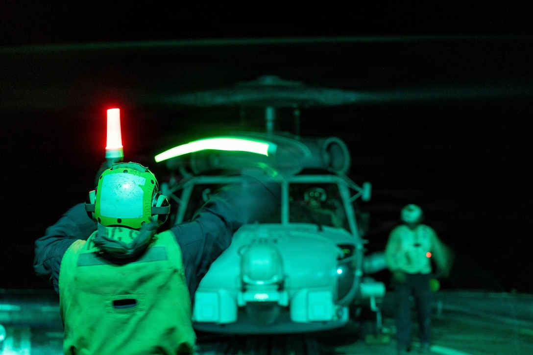A sailor waves a green baton while holding a red one in the opposite hand toward a sailor standing next to a helicopter aboard a ship in the dark.