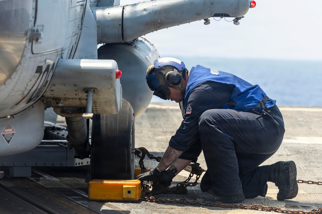 A sailor in a blue vest and helmet removes chains from a helicopter aboard a ship at sea during the day.