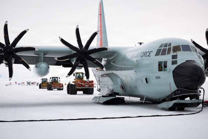 Three people driving large pieces of construction equipment with cargo attached drive toward a military aircraft sitting on a snowy tarmac. There is a long power cord attached to the aircraft.