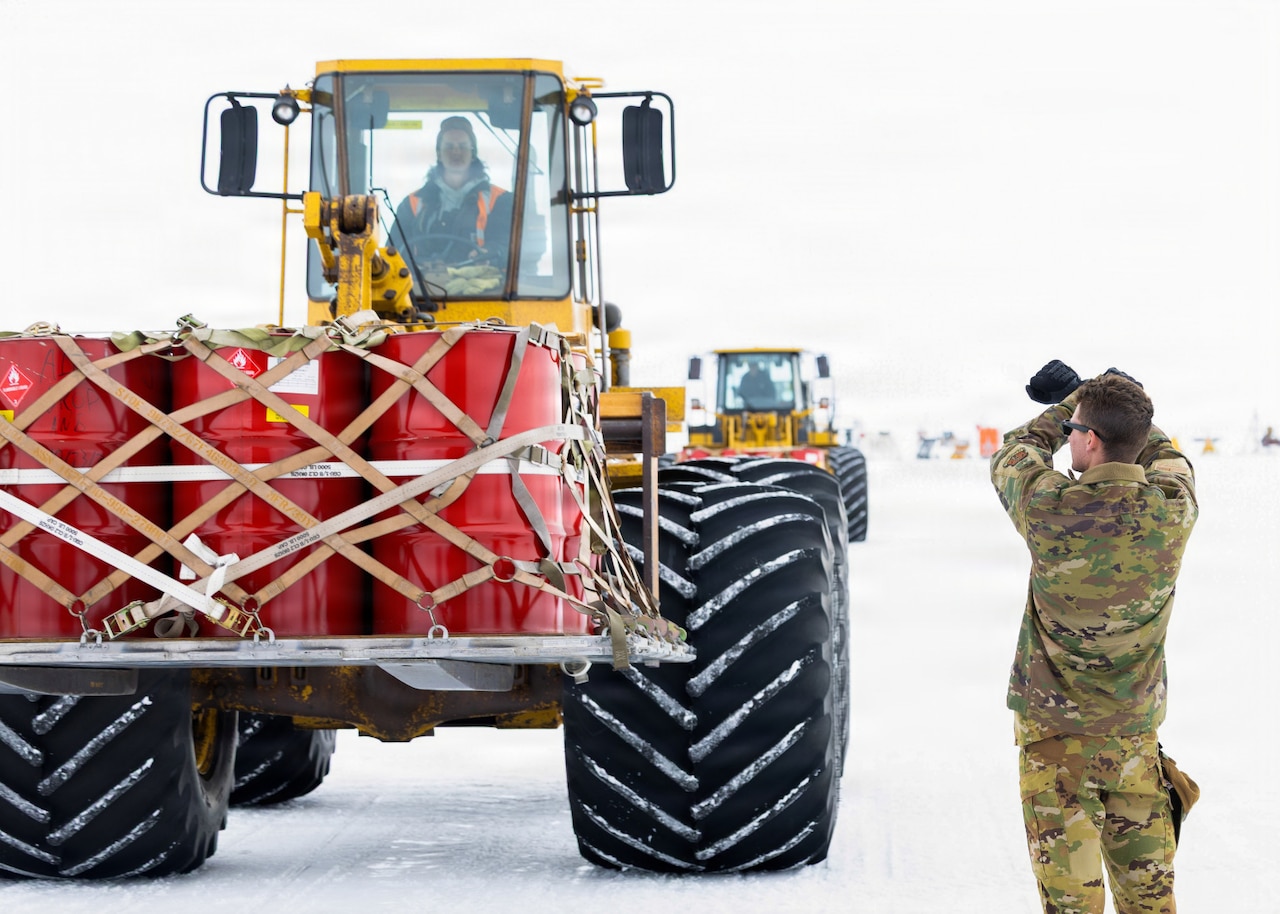 A man in a camouflage military uniform signals for a person driving a large piece of construction equipment with cargo attached to stop on a snowy tarmac. There is another piece of equipment with cargo in the background.