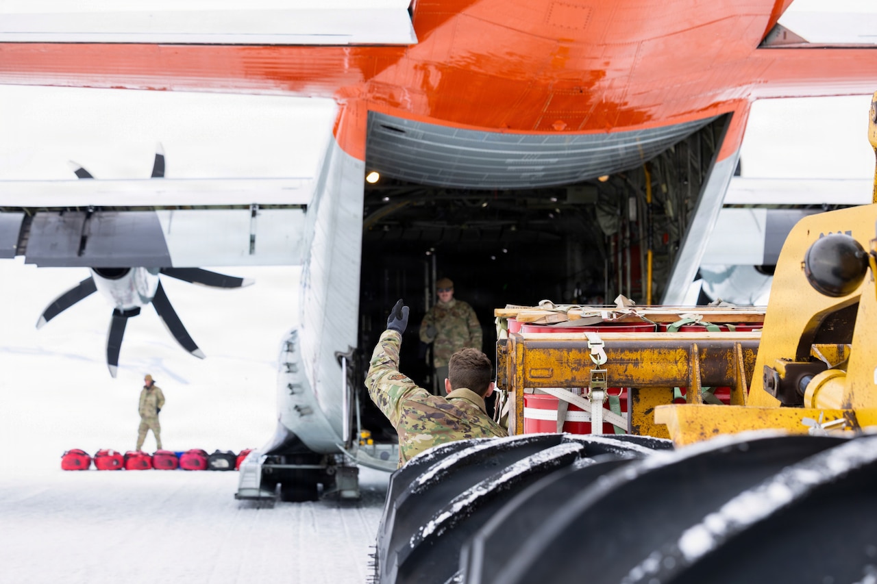 A man in a camouflage military uniform guides a loader with cargo into the back of a military aircraft sitting on a snowy tarmac. There is another man in similar attire standing in the aircraft.