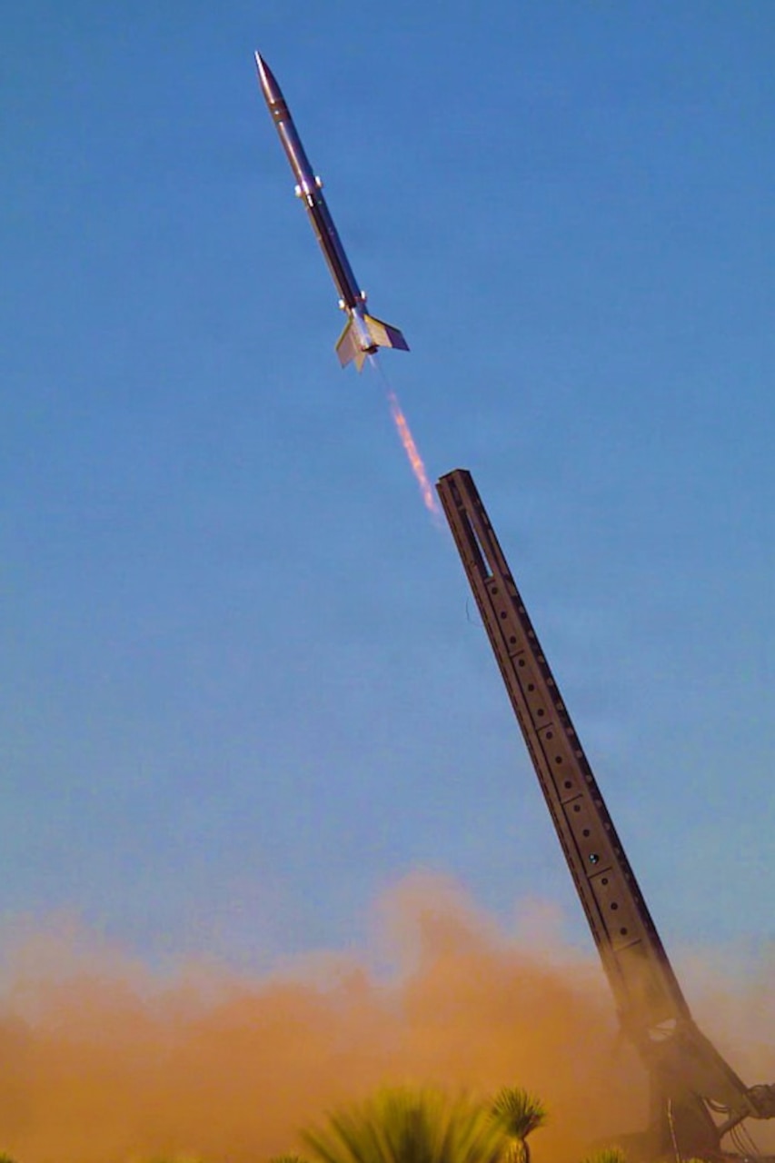 A missile launches into a clear blue sky, kicking up dust from the ground.