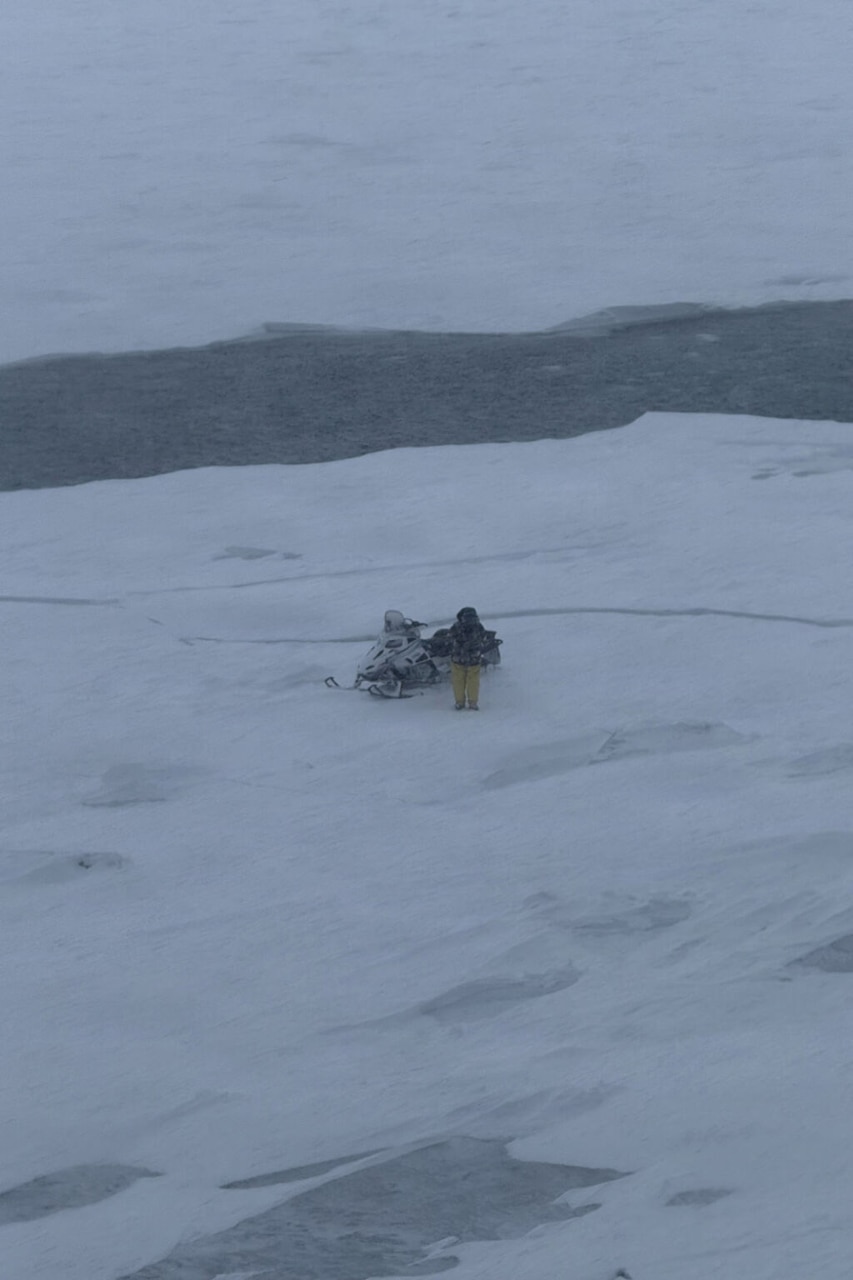 A man wearing protective winter gear stands next to a snowmobile on a floating piece of lake ice.