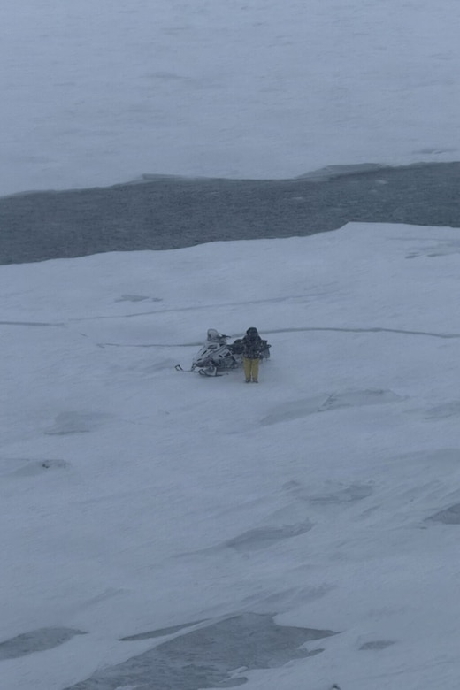 A man wearing protective winter gear stands next to a snowmobile on a floating piece of lake ice.