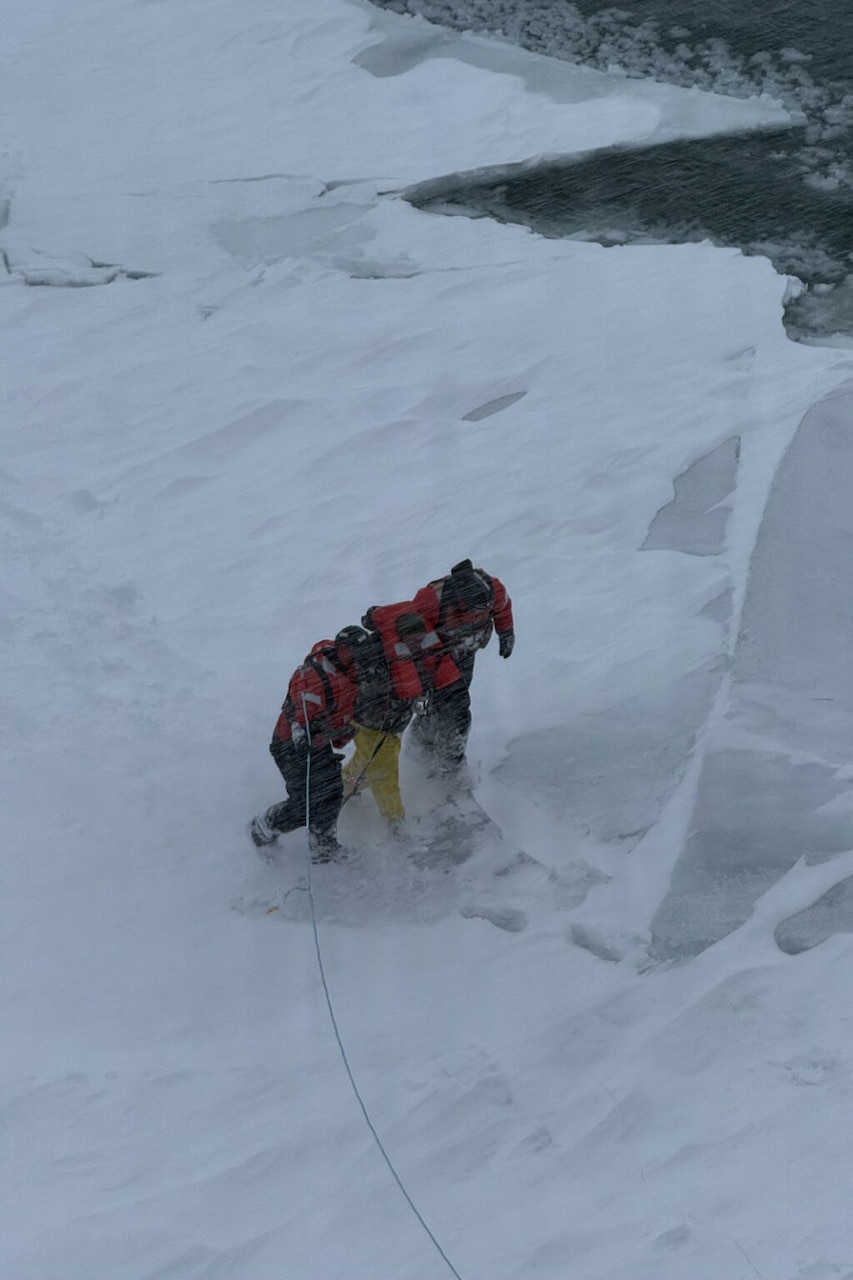 A man wearing protective winter gear is being assisted by two people in similar attire across ice during a rescue operation.