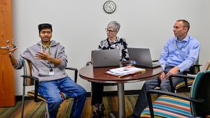Three people sit at a small table, two with laptops and one speaking while gesturing with his hands