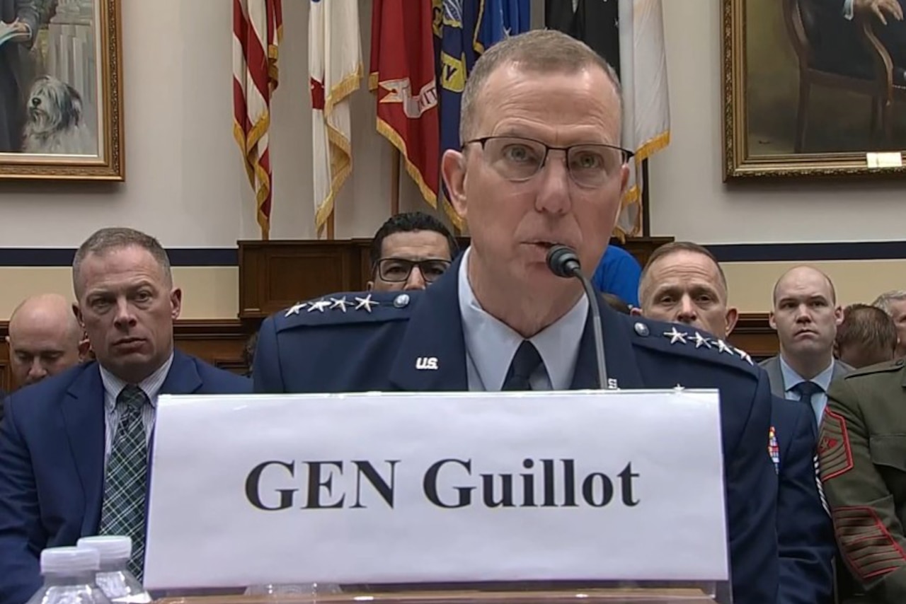 A man speaks into a microphone while sitting at a table in front of spectators.