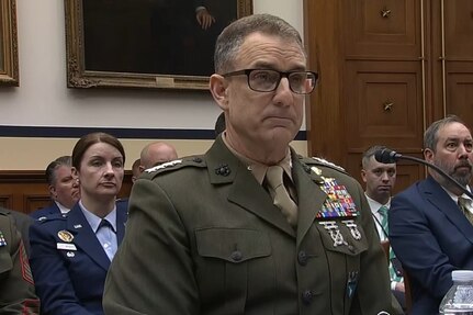 A man speaks into a microphone while sitting at a table in front of spectators.