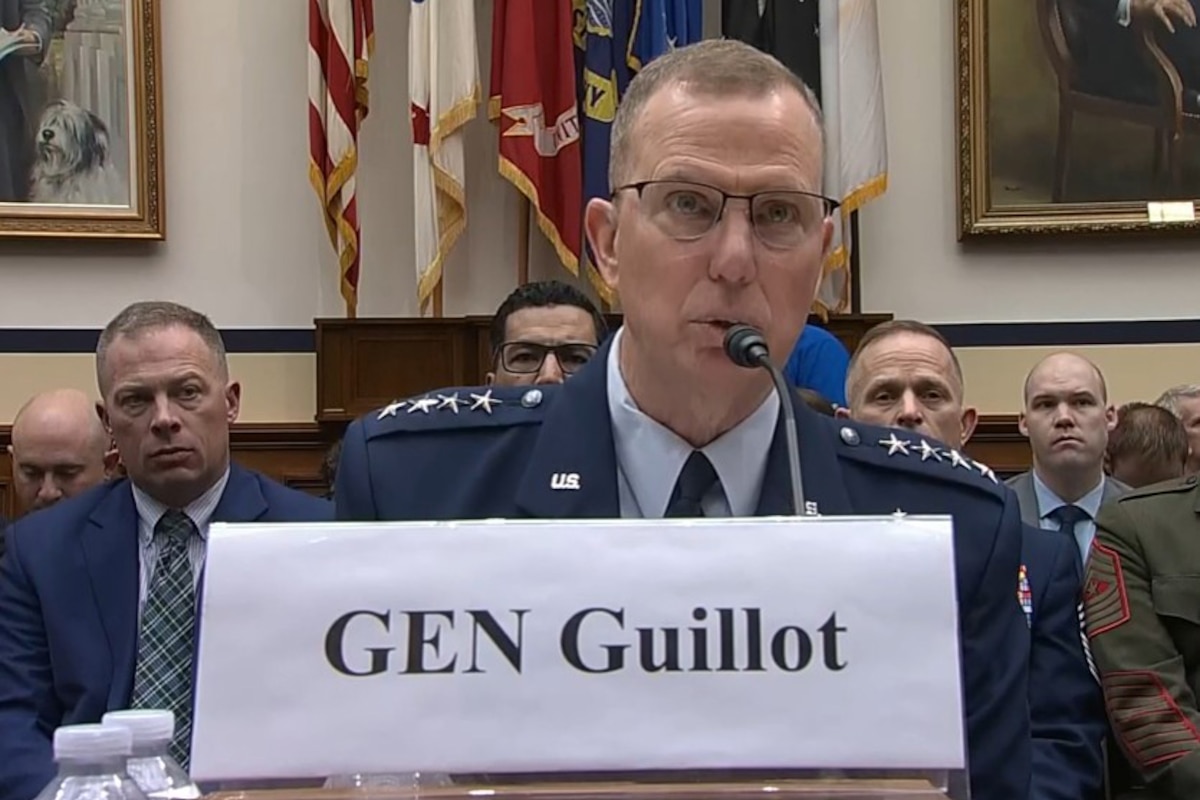 A man speaks into a microphone while sitting at a table in front of spectators.