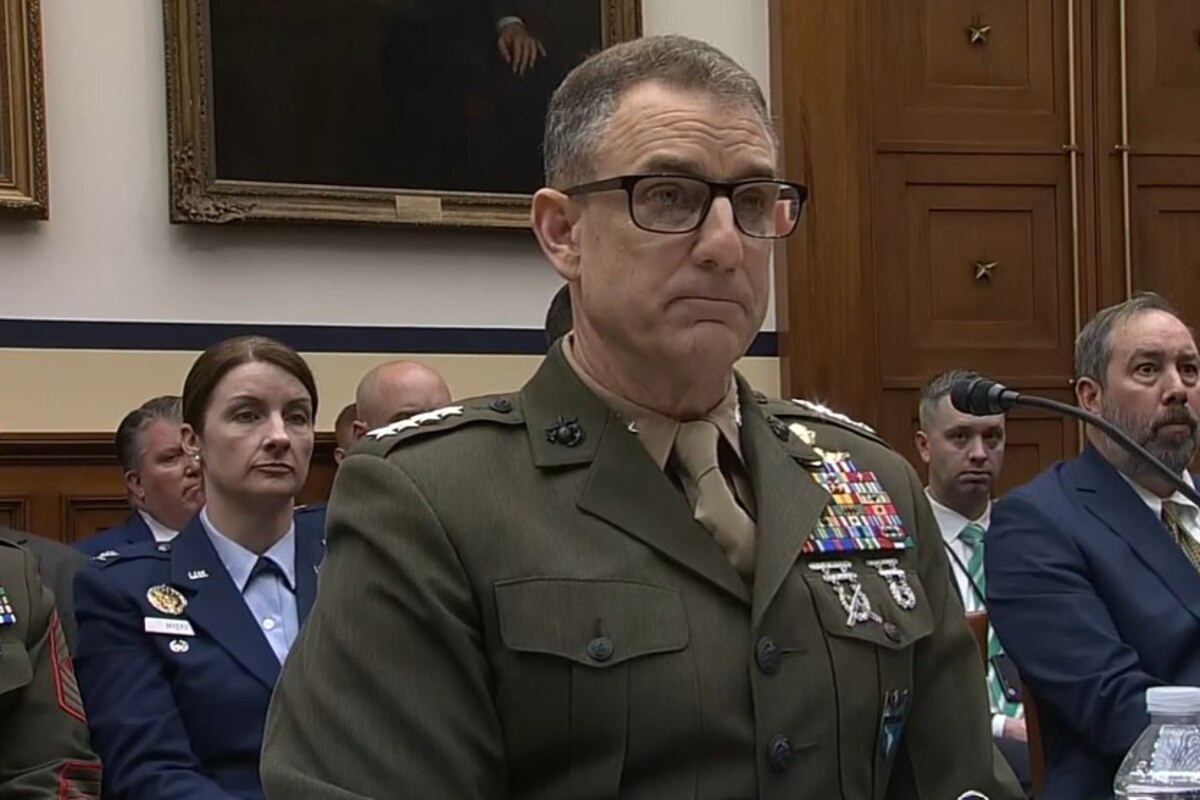 A man speaks into a microphone while sitting at a table in front of spectators.