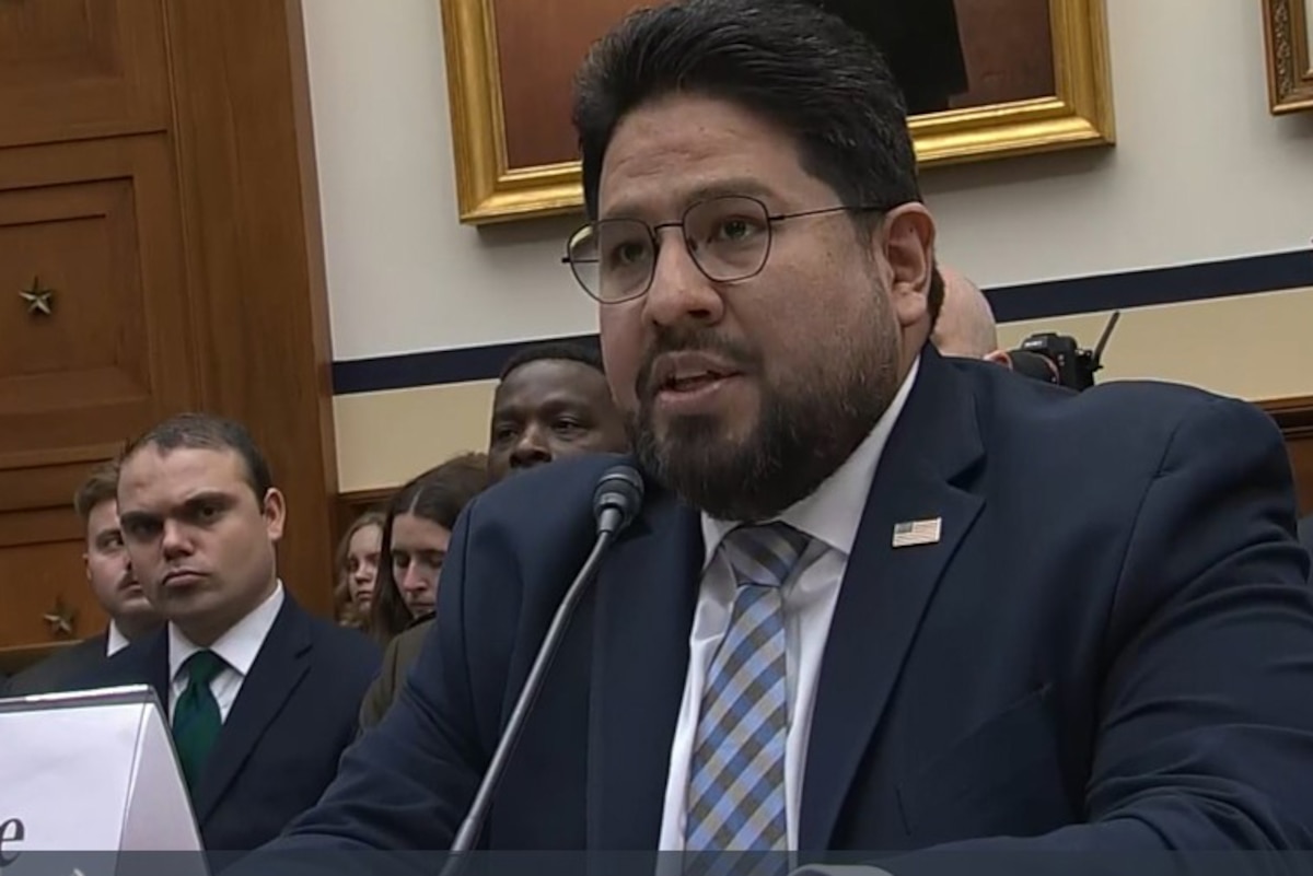 A man speaks into a microphone while sitting at a table in front of spectators.