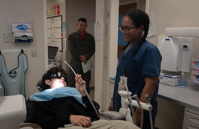 Aurelio (left), son of Tech. Sgt. Brian Cruz (center), 343rd Reconnaisance Squadron section chief of cyber support operations, holds a saliva ejector tool as a 55th Dental Squadron dental assistant stands by during Children's Dental Day at the Ehrling Bergquist clinic, March 6, 2026. The 55th DS provided a variety of dental services including flouride treatments and cleaning. (U.S. Air Force photo by Daniel Martinez)