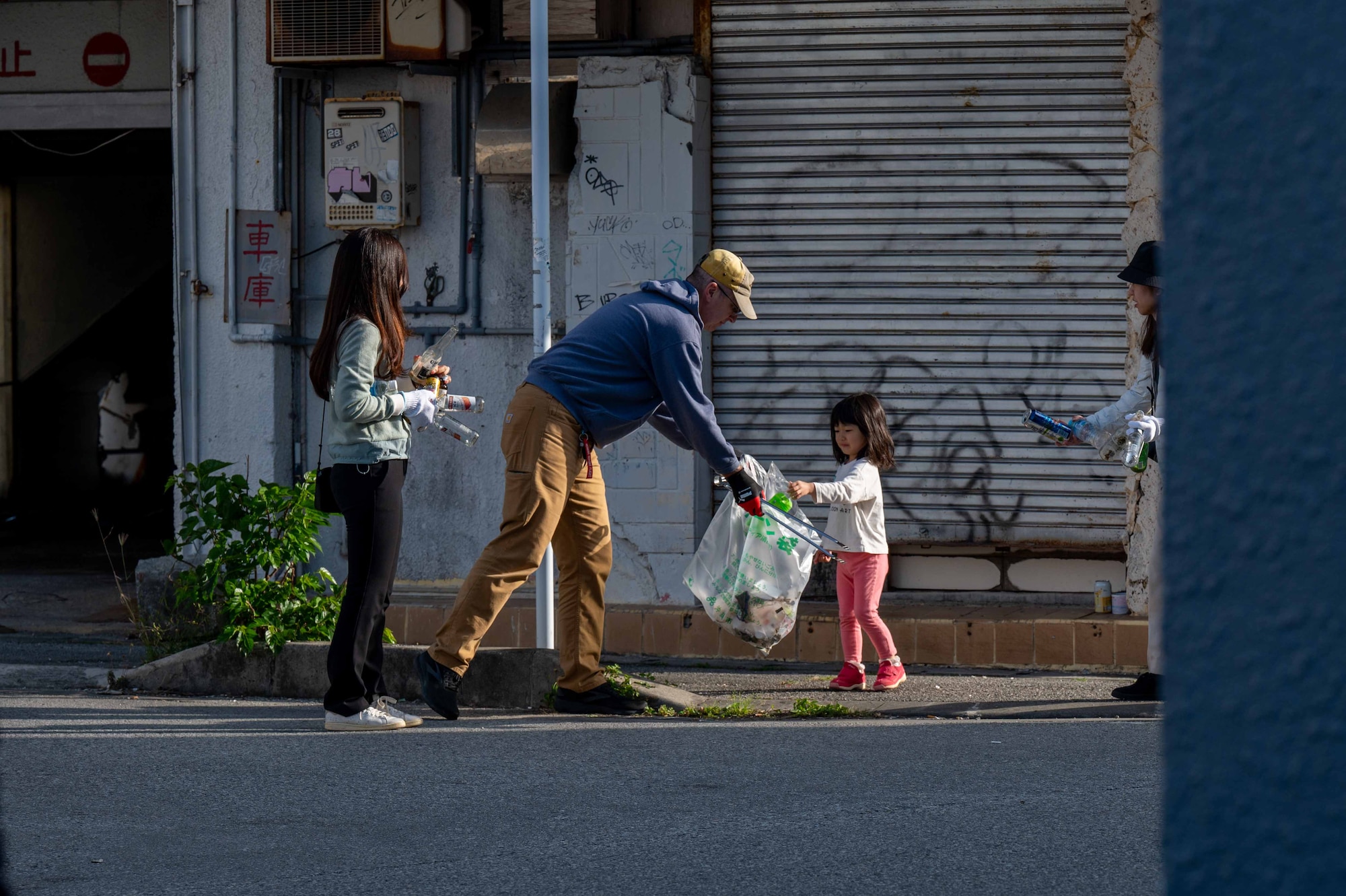 volunteers hold trash bags while walking down the street and one man holds out his trash bag for a little girl to place her trash in