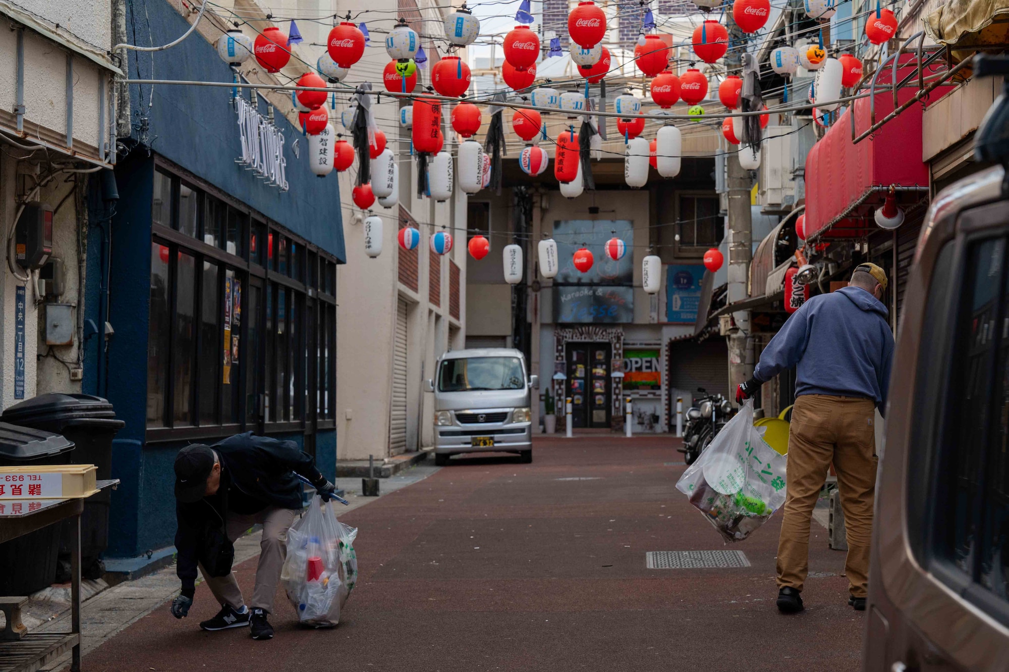 volunteers and civic leaders hold trash bags while walking down the street underneath red lanterns in a alley