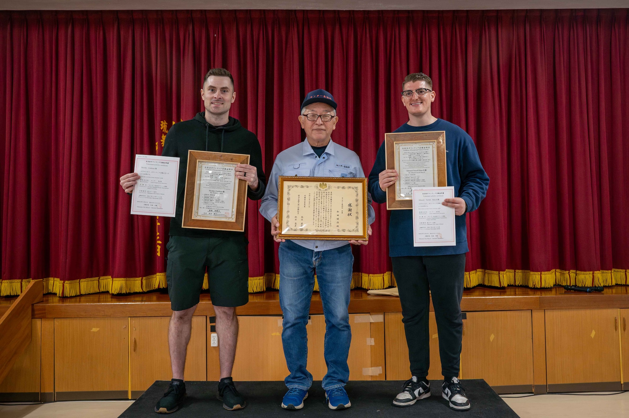 Three men stand on a podium holding certificates and plaques in front of the stage