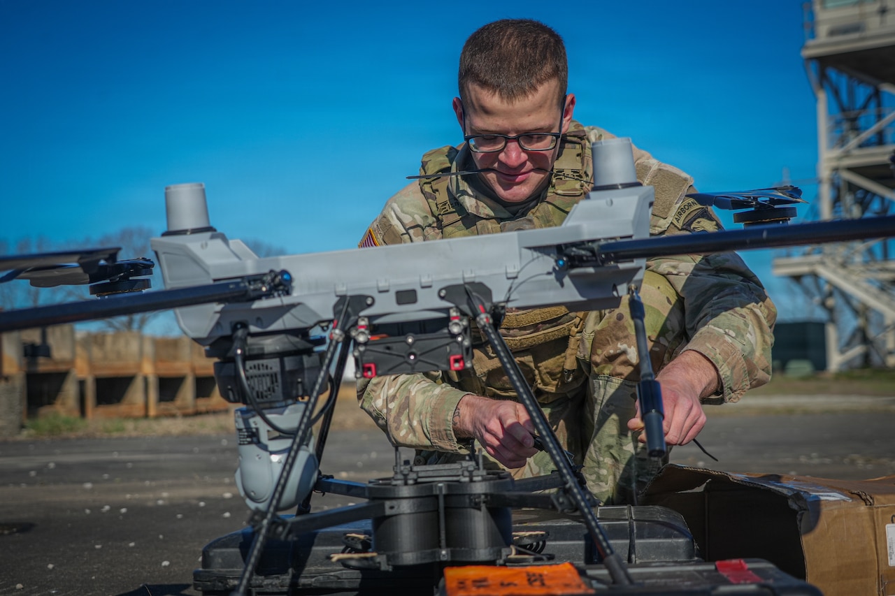 A man wearing a camouflage military uniform kneels as he works on a drone outside. There is equipment around the man, and a large tower is in the background.