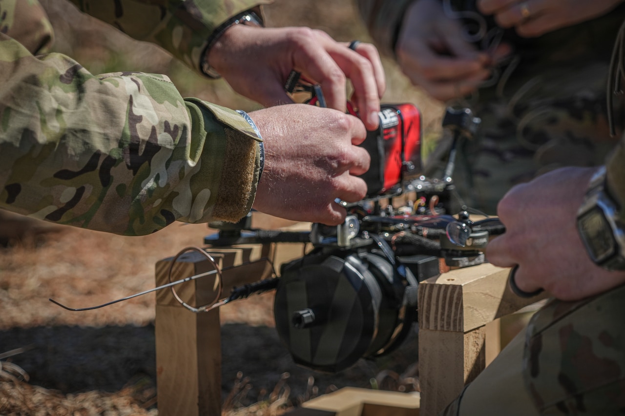 A close-up image of a person's hands as they work on a drone that is sitting on two pieces of wood outside. There are two other people kneeling around the drone.