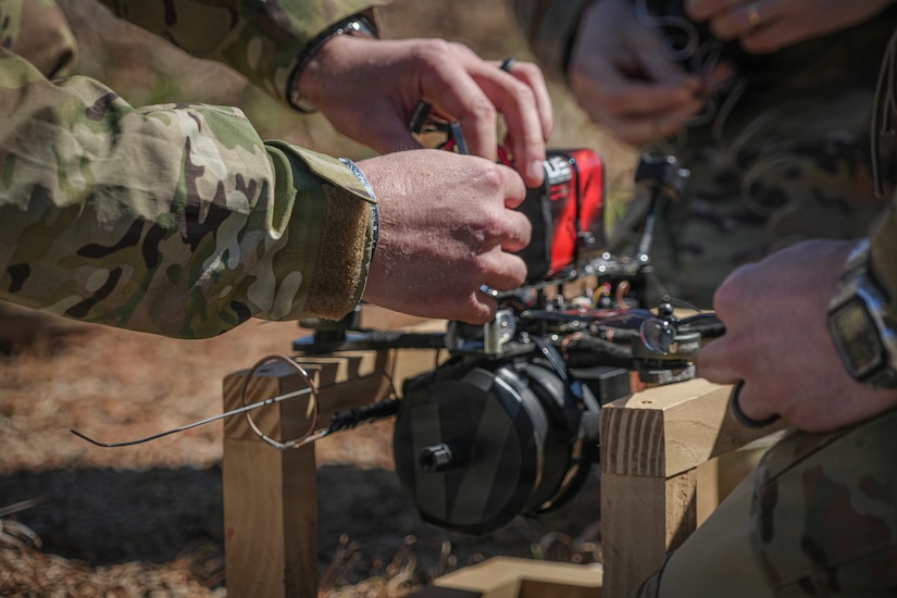 A close-up image of a person's hands as they work on a drone that is sitting on two pieces of wood outside. There are two other people kneeling around the drone.