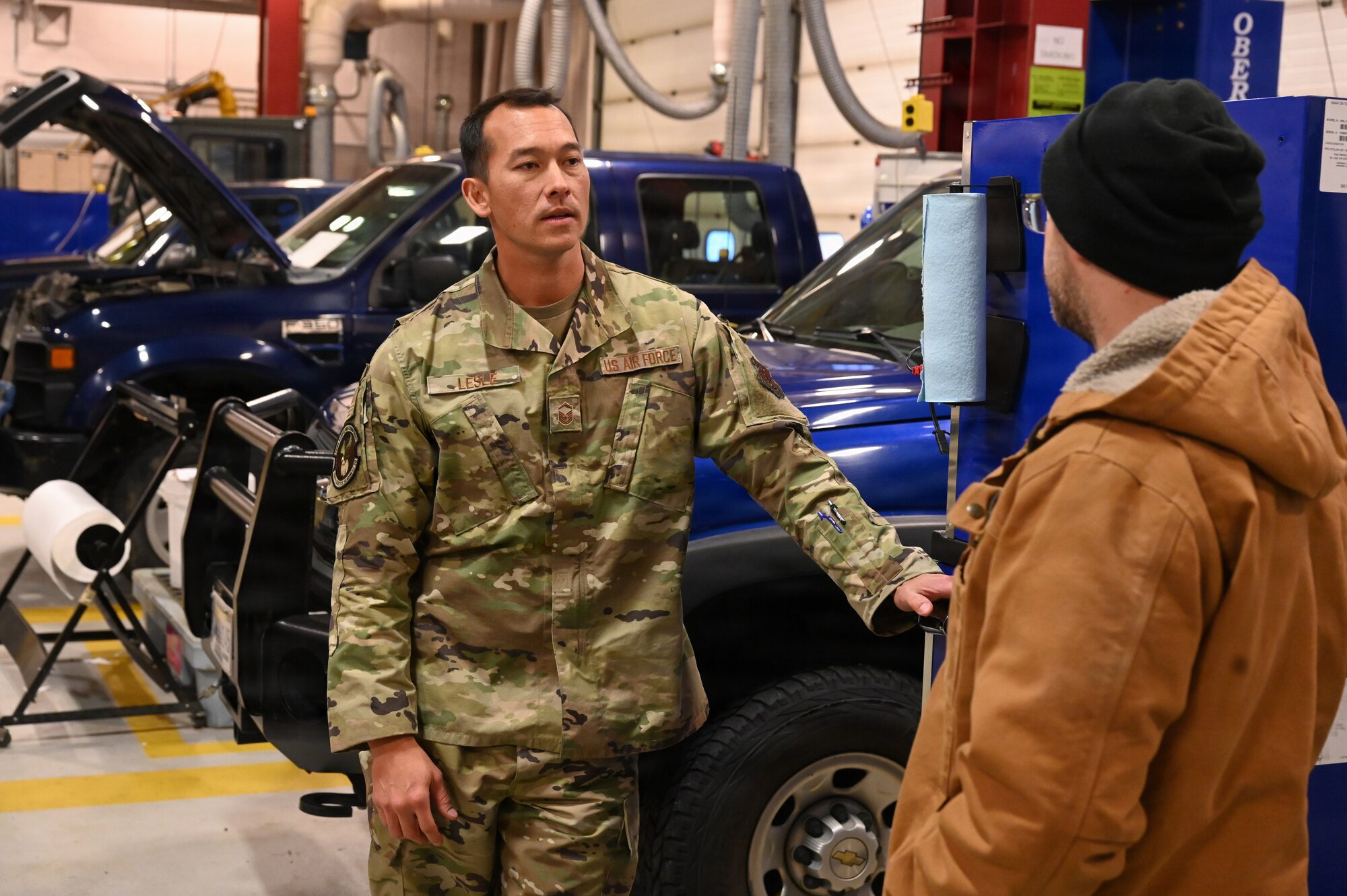 A U.S. Air Force Airman leans near a truck, talking with a man in a work jacket.