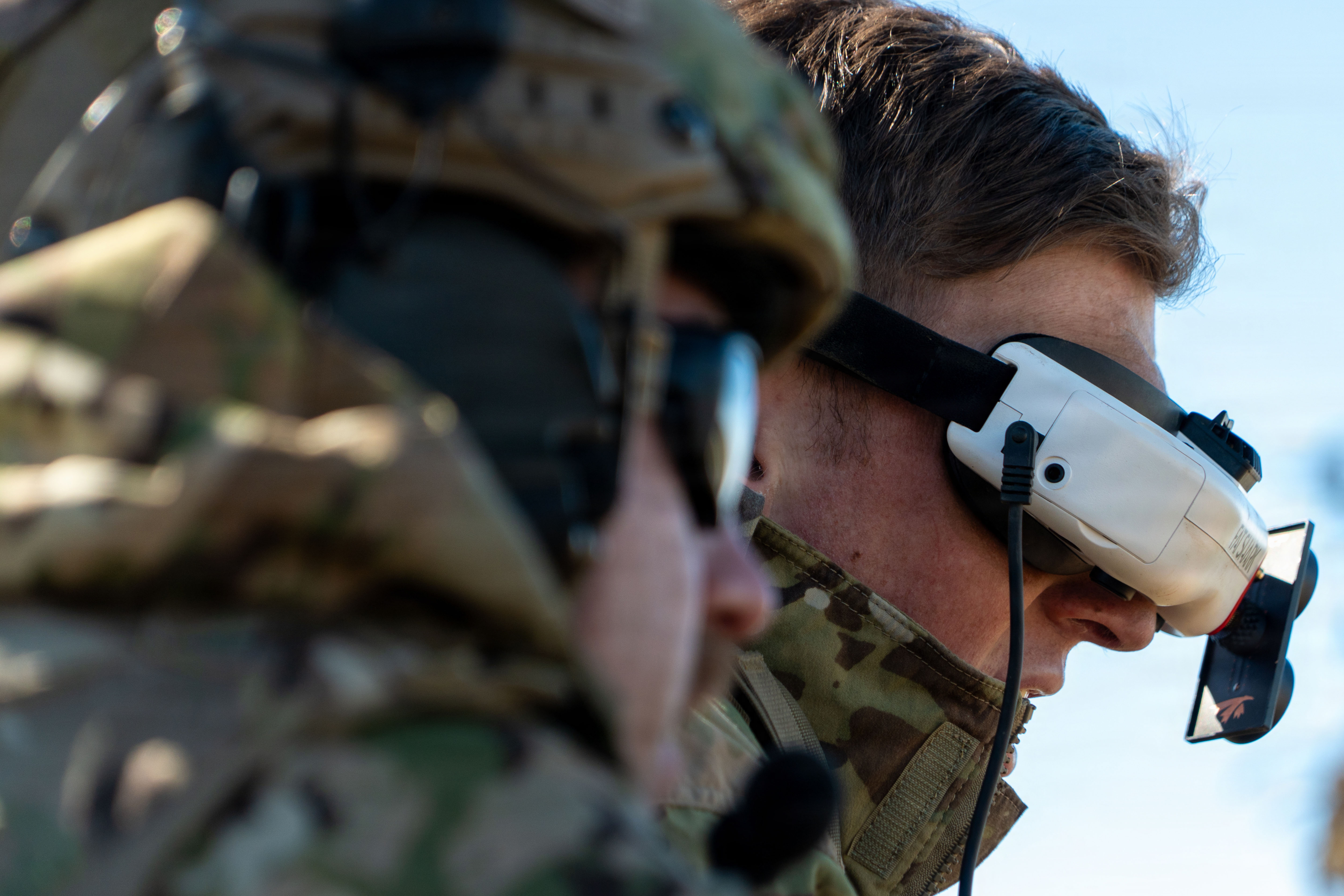A close-up image of a man wearing a camouflage military uniform with a drone headset on. There is another man in similar attire in the foreground. 