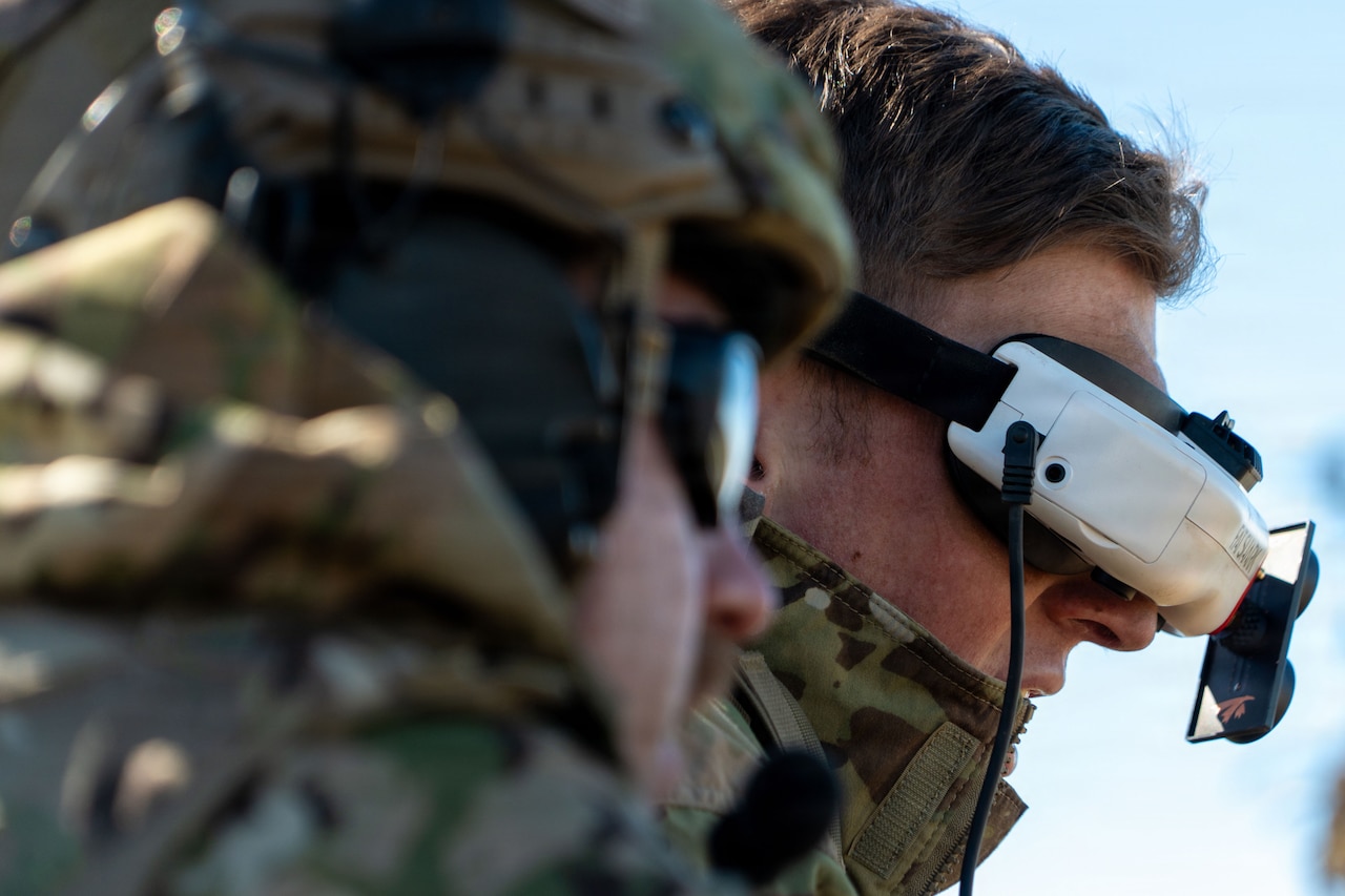 A close-up image of a man wearing a camouflage military uniform with a drone headset on. There is another man in similar attire in the foreground.