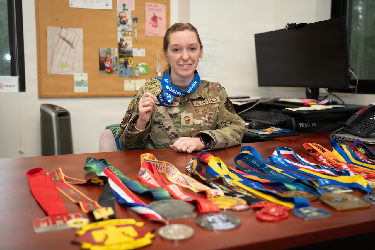 A woman wearing a camouflage military uniform sits at a table with multiple racing medals and awards. The woman is holding up one of the medals as it hangs around her neck.