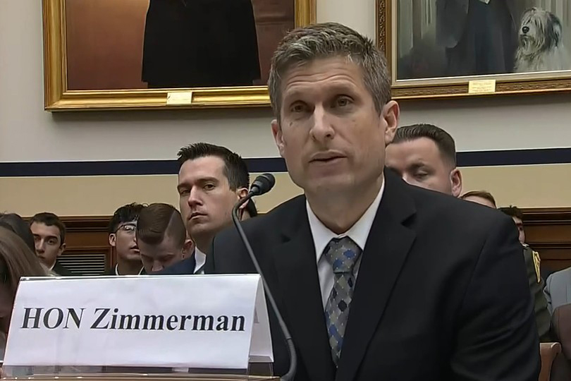 A man in a suit, seated at a table, speaks into a microphone.