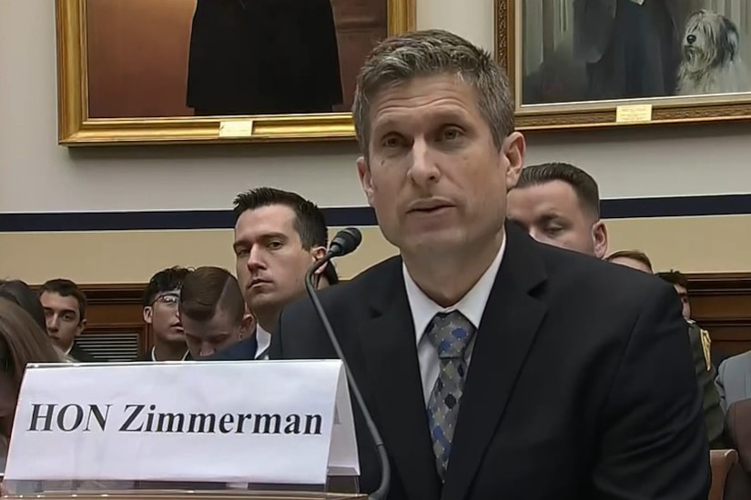 A man in a suit, seated at a table, speaks into a microphone.