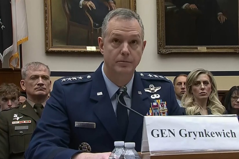 A man in a dress uniform, seated at a table, speaks into a microphone.