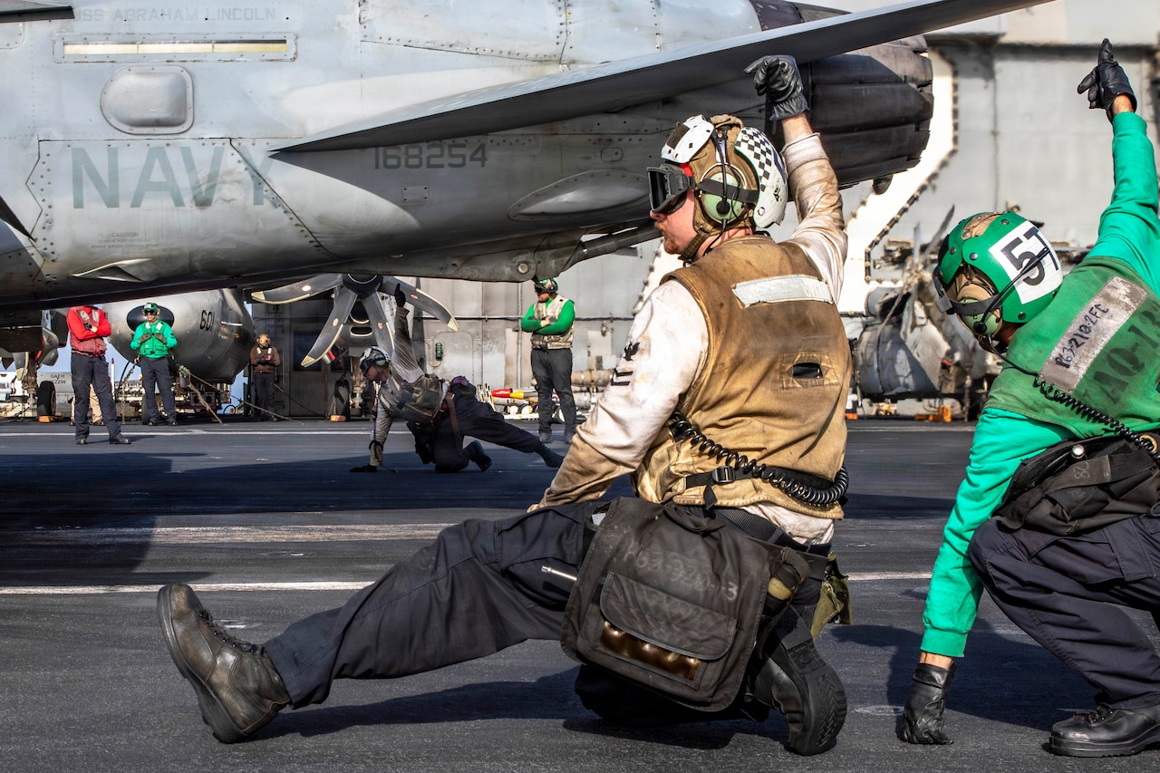 Sailors leaning on the surface of a ship give a thumbs-up near a partially visible aircraft during the day.