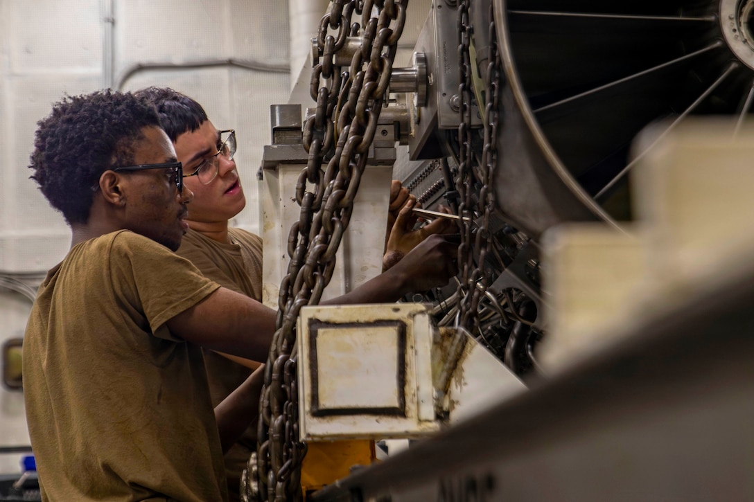 Sailors in brown shirts use tools to work on an engine secured by chains in a maintenance shop.