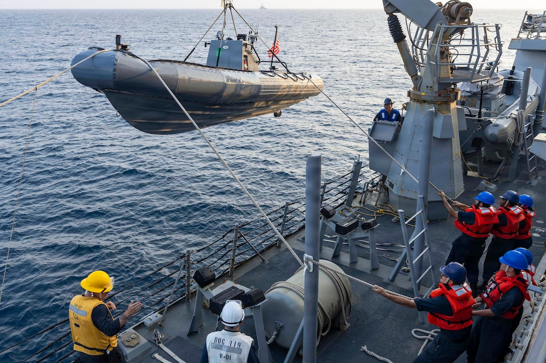 A group of sailors in life vests lower a small boat off the side of a ship as three fellow sailors watch during the day.