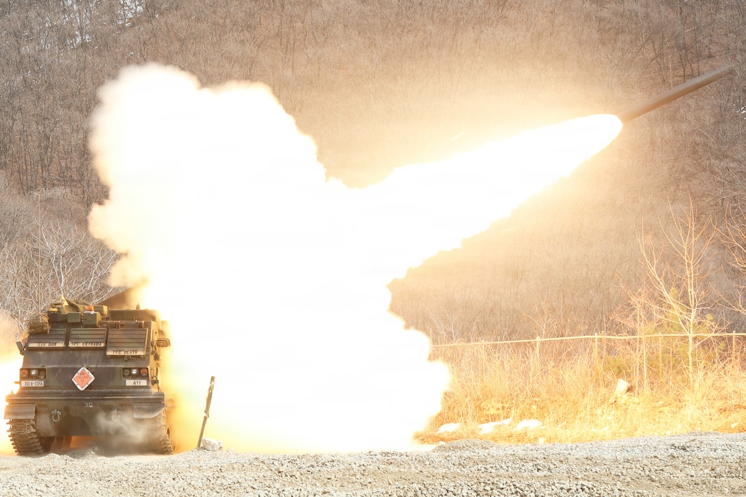 A military vehicle fires a rocket in a field, creating a large, bright yellowish blast.