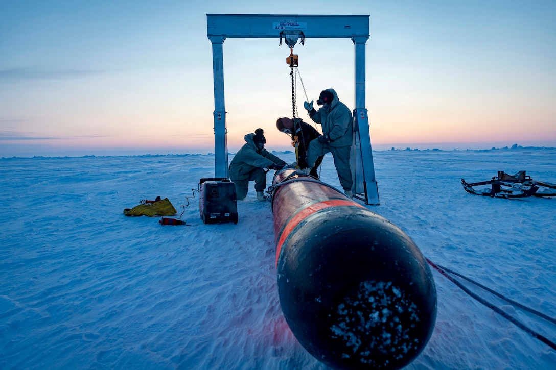 Three Navy personnel in winter gear tie cords around a black torpedo on a snowy surface as pinkish hues hover above the horizon.