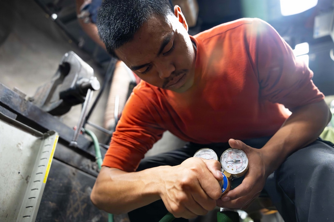A sailor in an orange shirt bends over to check the pressure of an oxygen tank in maintenance room.