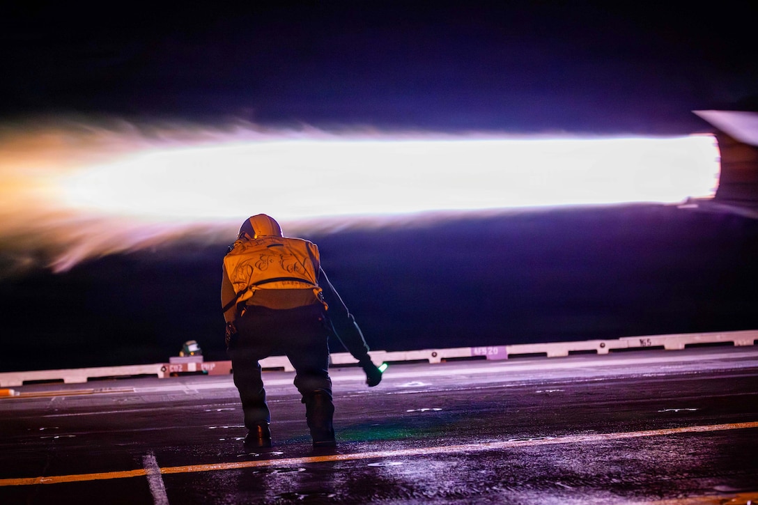 A sailor in a yellow vest bends over on a ship as a partially visible aircraft takes off in the dark, creating a bright blast.