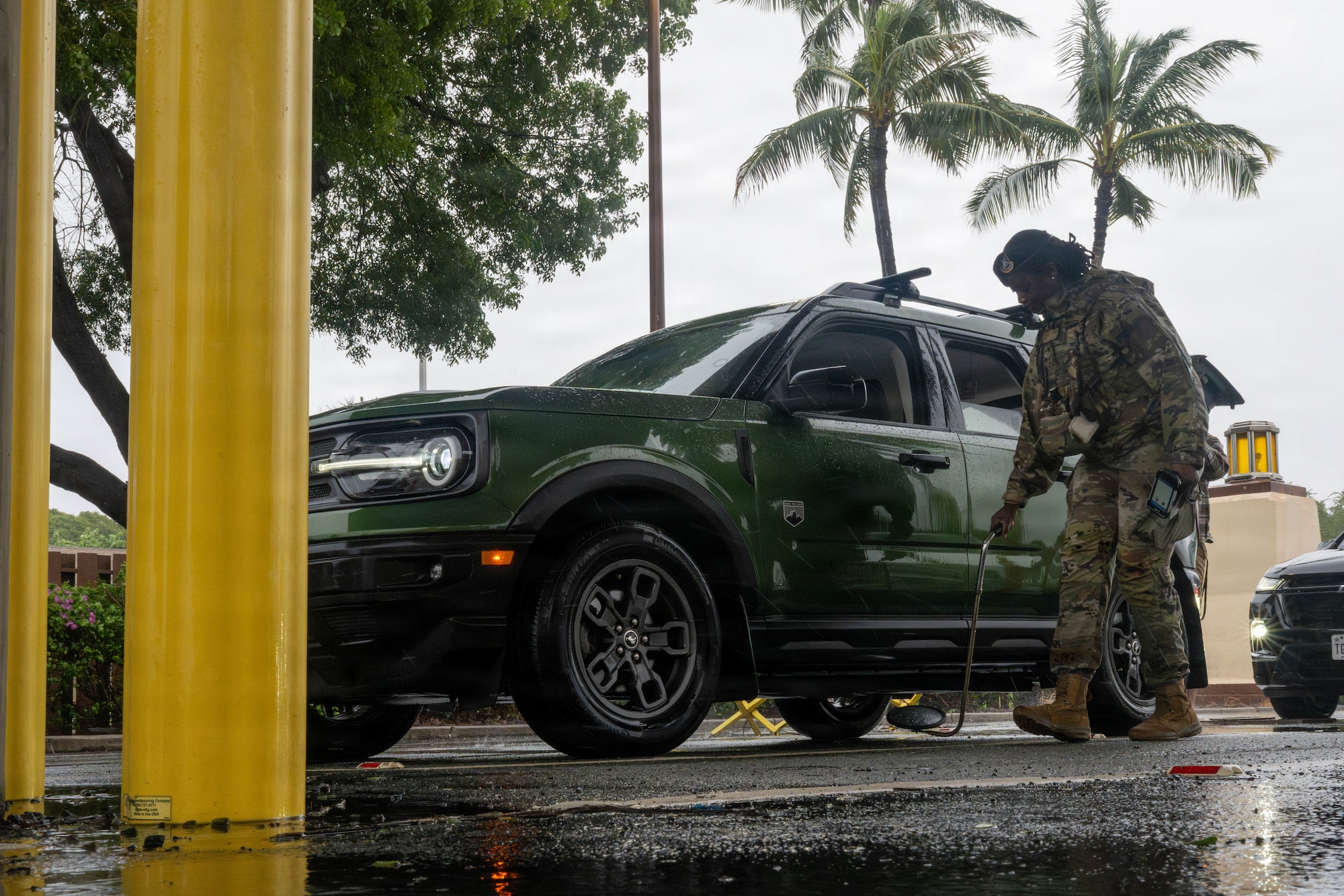 An Airman uses a pole with a mirror attached at the end to view the underside of a vehicle.