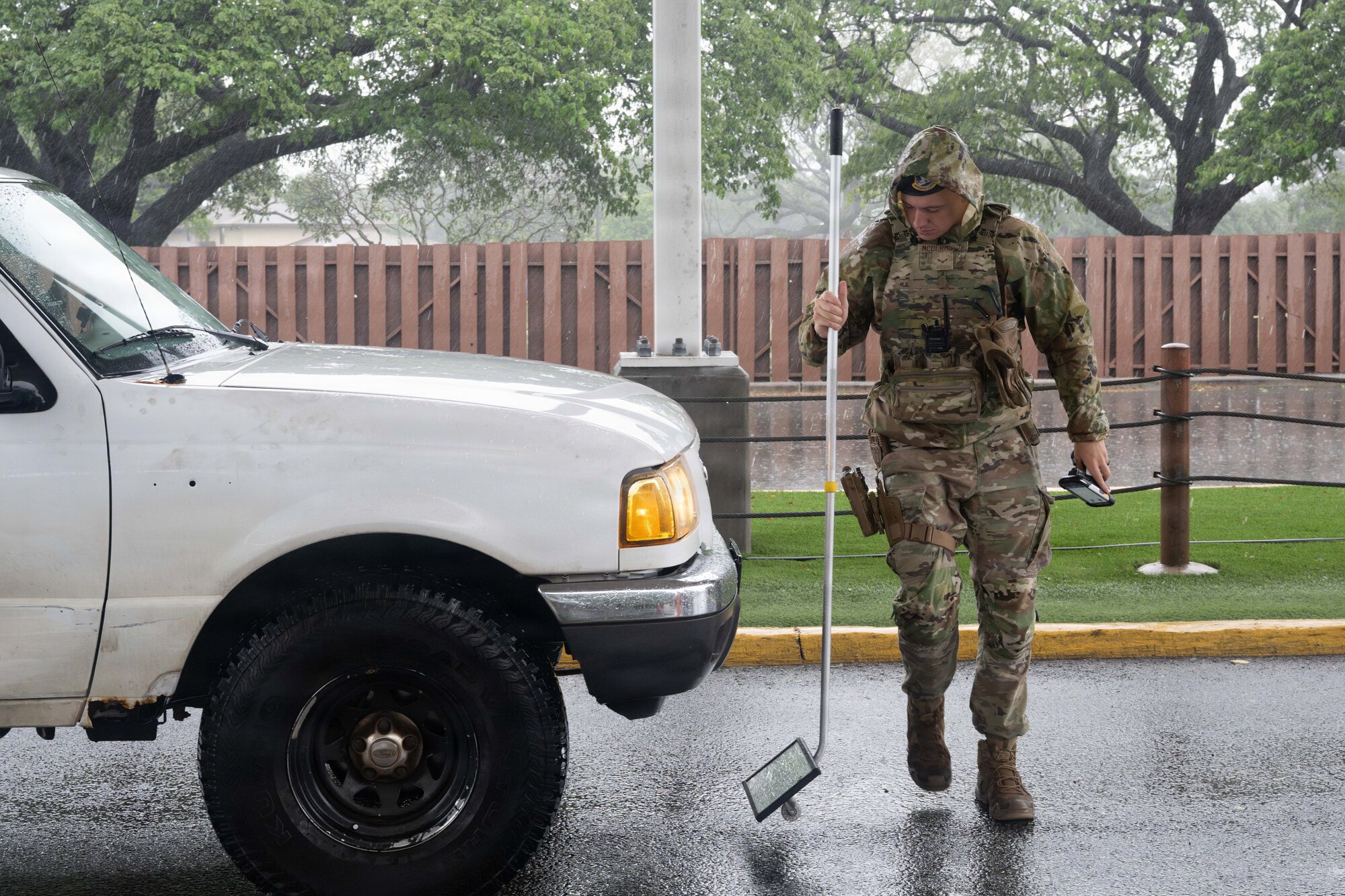 Airman uses a long pole with a mirror attached at the end to view the underside of a vehicle as they walk around it.