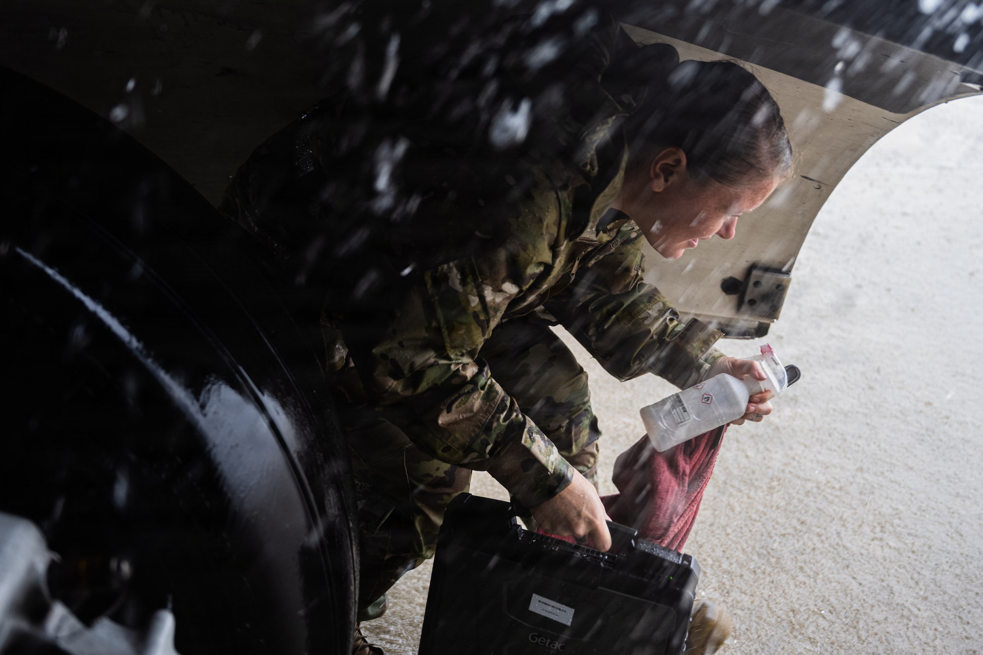 An Airman squats while exiting the wheel well of an aircraft while holding multiple tools as rain pours down.